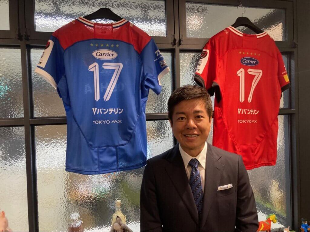 自身が代表を務める株式会社17(ジュウナナ)のオフィスに飾られた東芝ブレイブルーパス東京のユニホームの前に立つ松尾卓哉氏(原田写す)