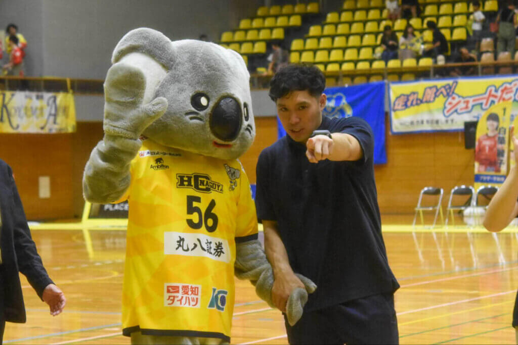 チームマスコットのコロちゃんとともに、開幕戦勝利の挨拶をする藤本純季コーチ。2025年9月、名古屋市枇杷島スポーツセンターで（久保写す、以下すべて）