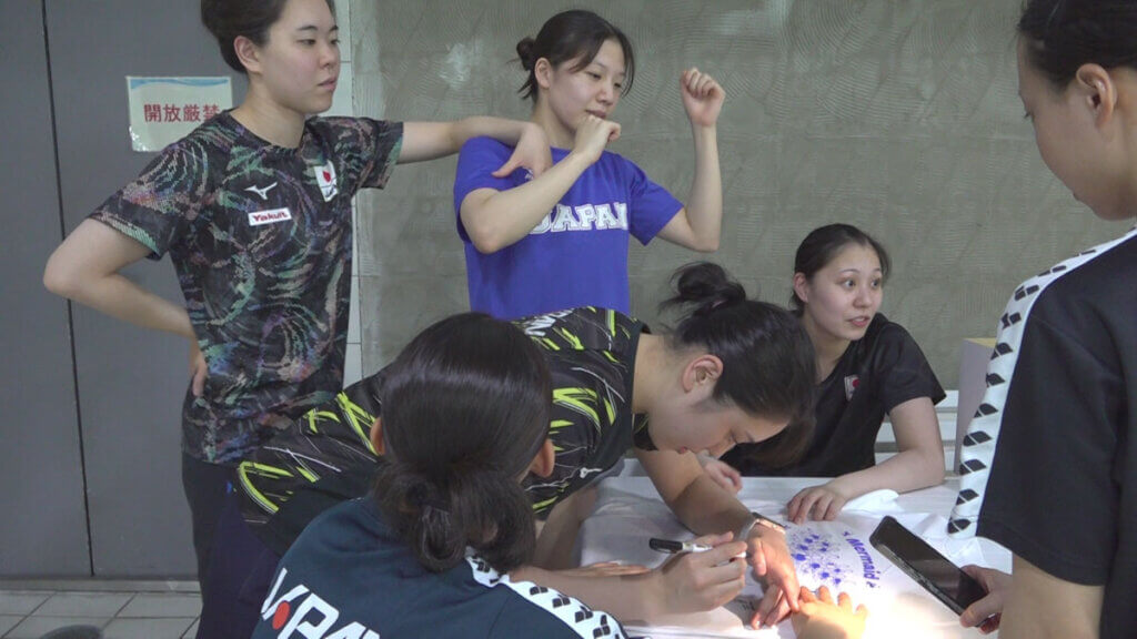 クラファンのお返しでサインをする選手の横で1人踊る小林選手(青いTシャツ)